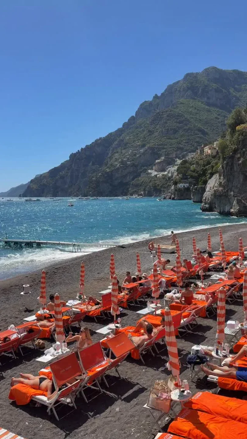 Sunbathers relax on orange loungers at Arienzo Beach Club, Amalfi Coast, with turquoise waters and dramatic cliffs.