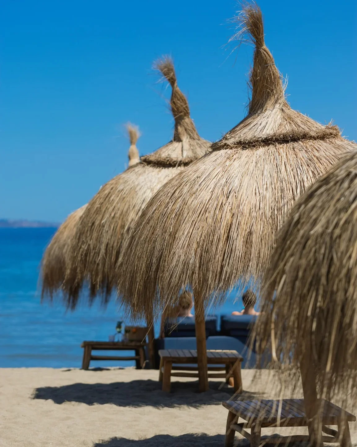 Banus Beach club exterior with thatched umbrellas and lounge chairs overlooking the turquoise sea under a clear blue sky.