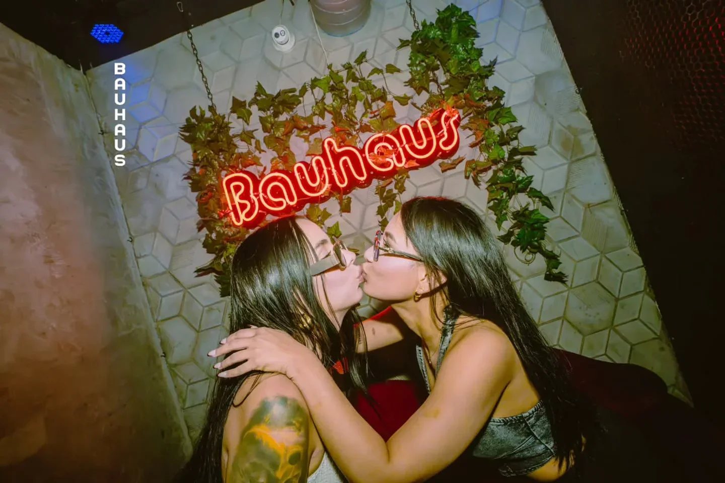 Two women kissing at Bauhaus nightclub in Houston, with a neon sign in the background, nightlife scene.
