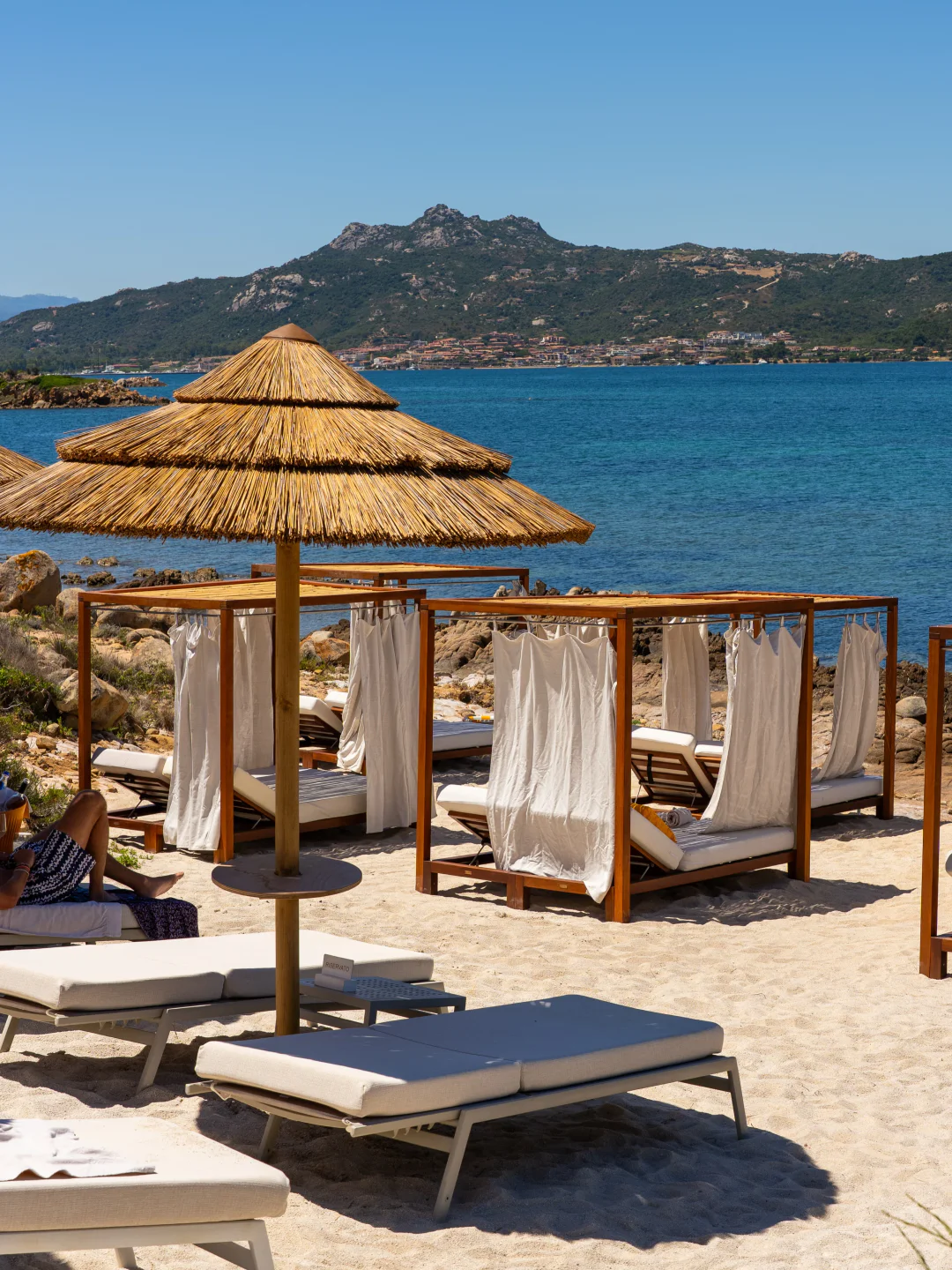 Cone Club beach club in Porto Cervo with cabanas and loungers on the sand overlooking the blue sea and hills.