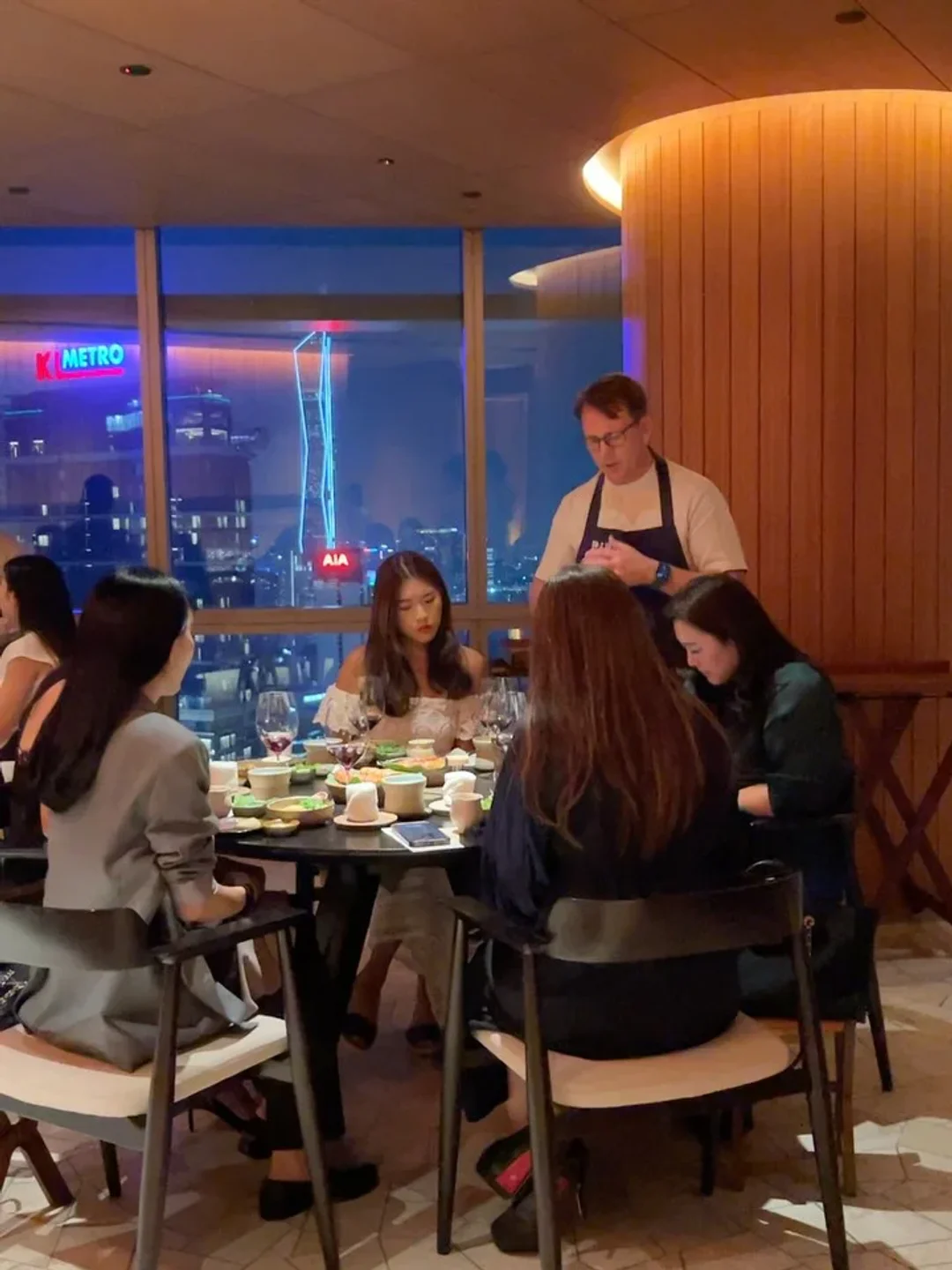 Dining scene at Dewakan restaurant in Kuala Lumpur with city views through the window and a server attending a table.