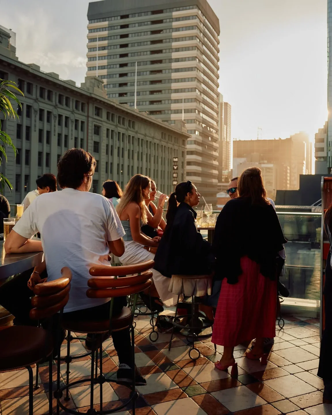 People enjoy drinks at HER Rooftop in Melbourne with city buildings in the background during sunset