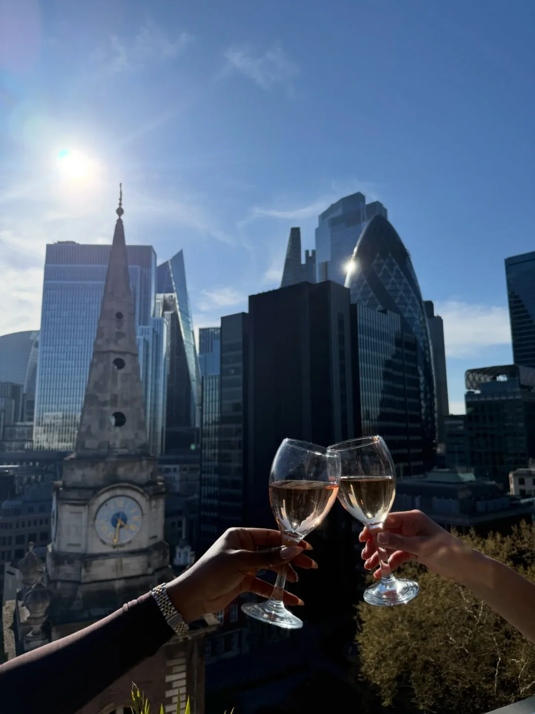 Two glasses of wine clinking on the rooftop of Jin Bo Law, London, with skyscrapers in the background.