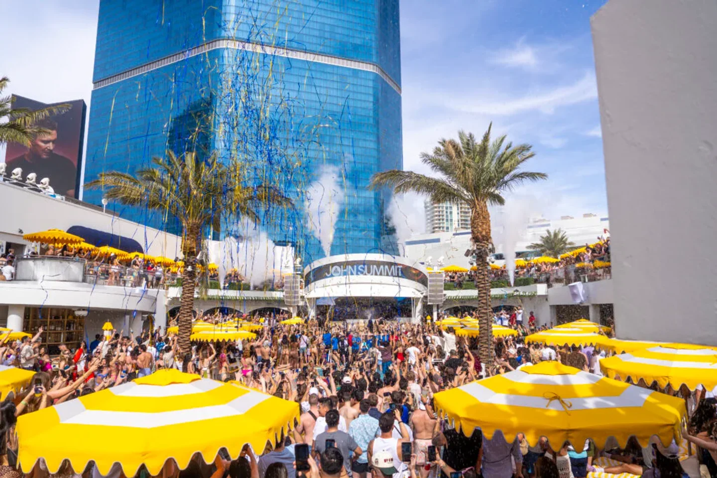 Confetti rains down on a lively crowd at LIV Beach Las Vegas during a vibrant daytime party with palm trees and a blue skyscraper.