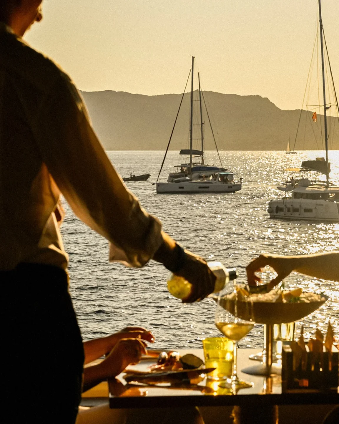 Dine al fresco at Luciano's Porto Cervo with a sunset view of sailboats and coastal mountains.