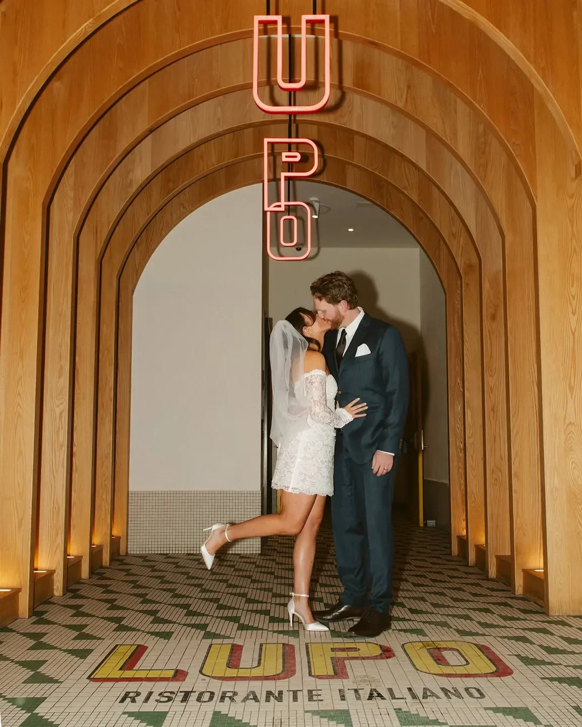 Newlyweds kiss at LUPO Ristorante Italiano in Banff, under a neon sign and wooden archway.
