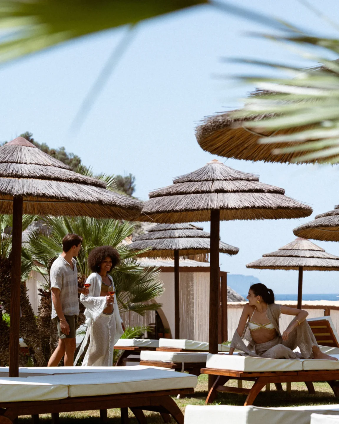 People enjoying drinks at Maya Beach Experience, Amalfi Coast, under straw umbrellas and palm trees.