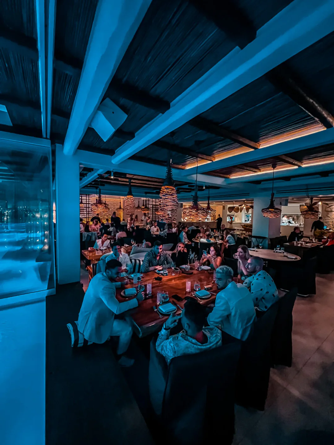 Meraki Mykonos restaurant interior features stylish decor, ambient lighting, and patrons enjoying dinner at wooden tables.