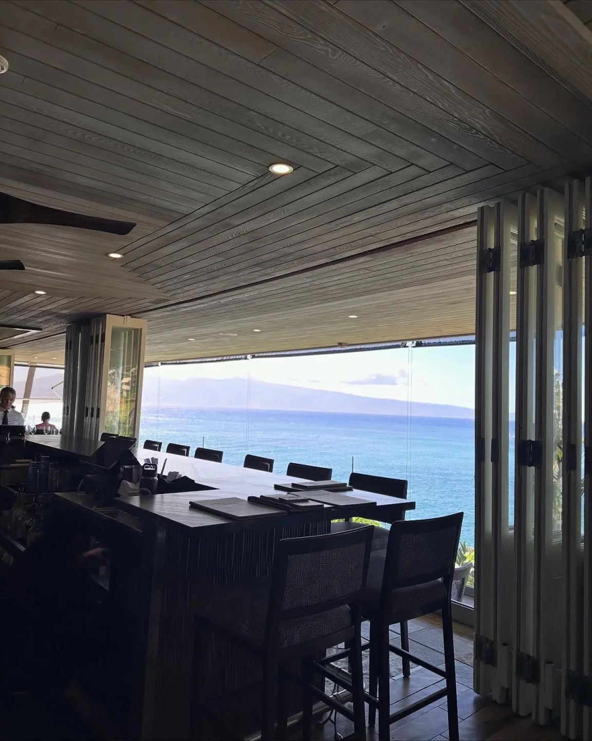 Stunning ocean view from Merriman's Kapalua restaurant in Maui. Dining tables overlooking the Pacific Ocean and landscape.
