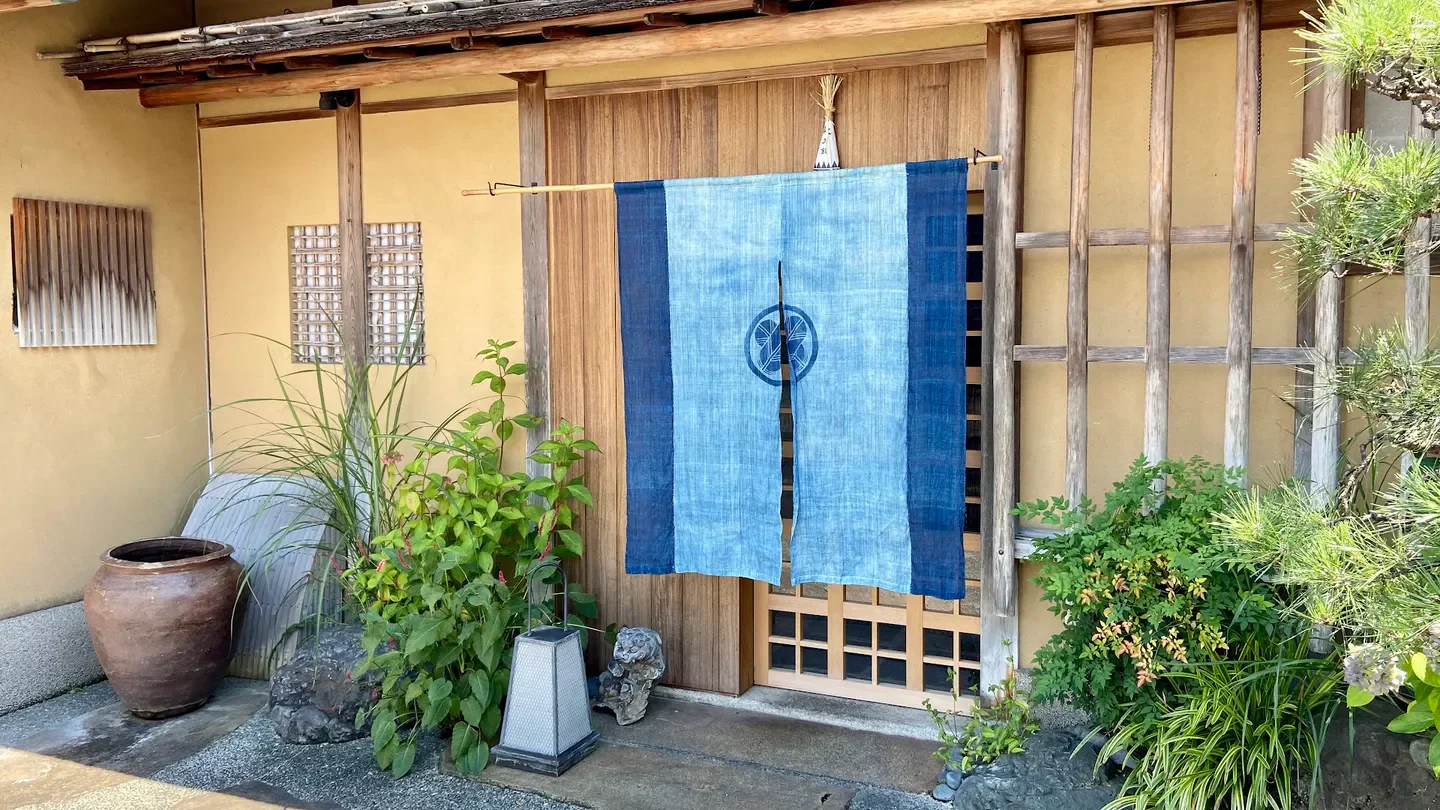 Entrance of Okina restaurant in Kyoto, featuring traditional blue noren and wooden architecture.