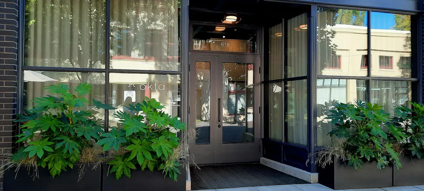 Entrance of ōkta restaurant in Willamette Valley, with large windows and lush greenery.