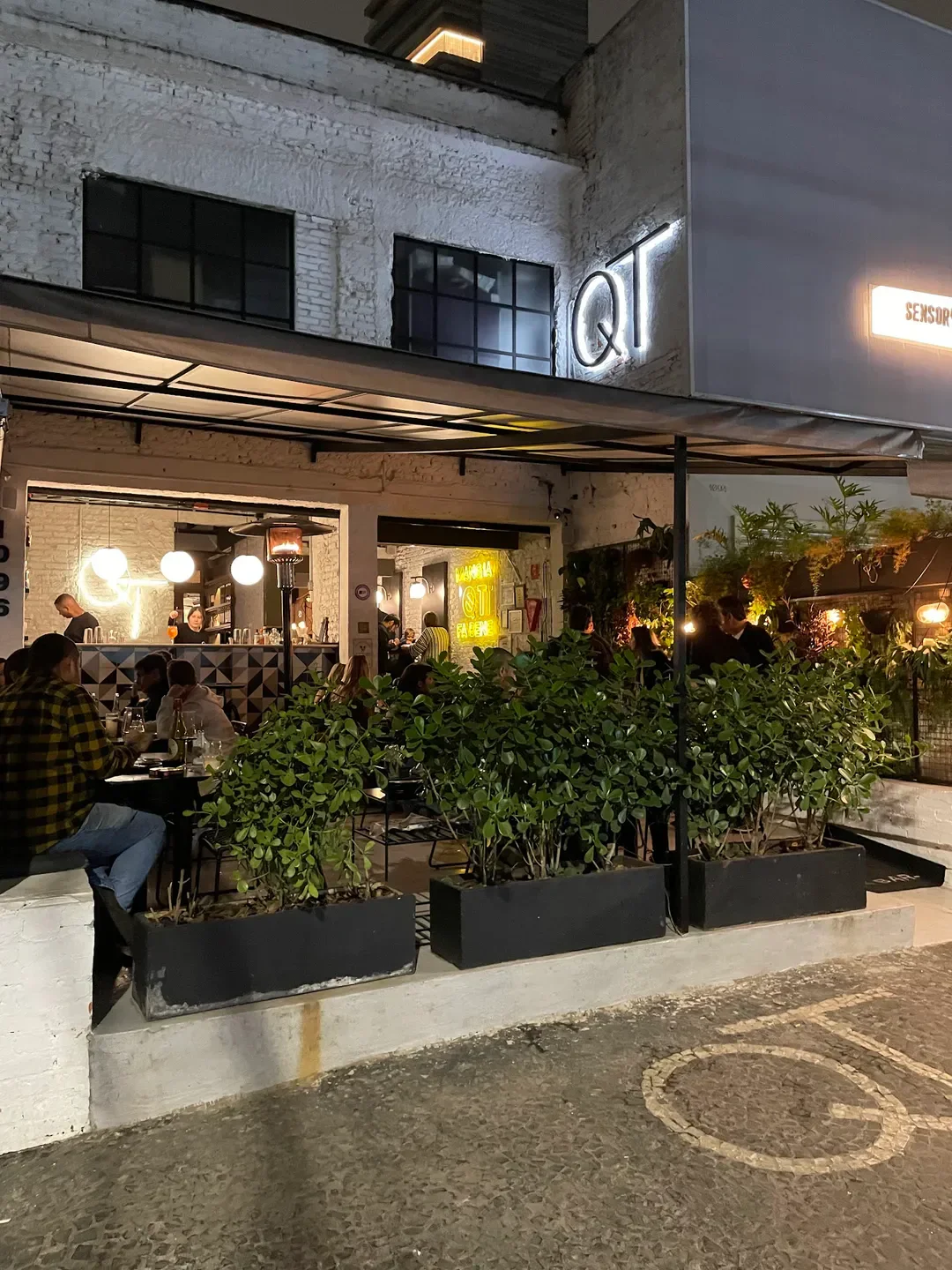 Exterior of QT Pizza Bar in Sao Paulo with outdoor seating and neon sign.