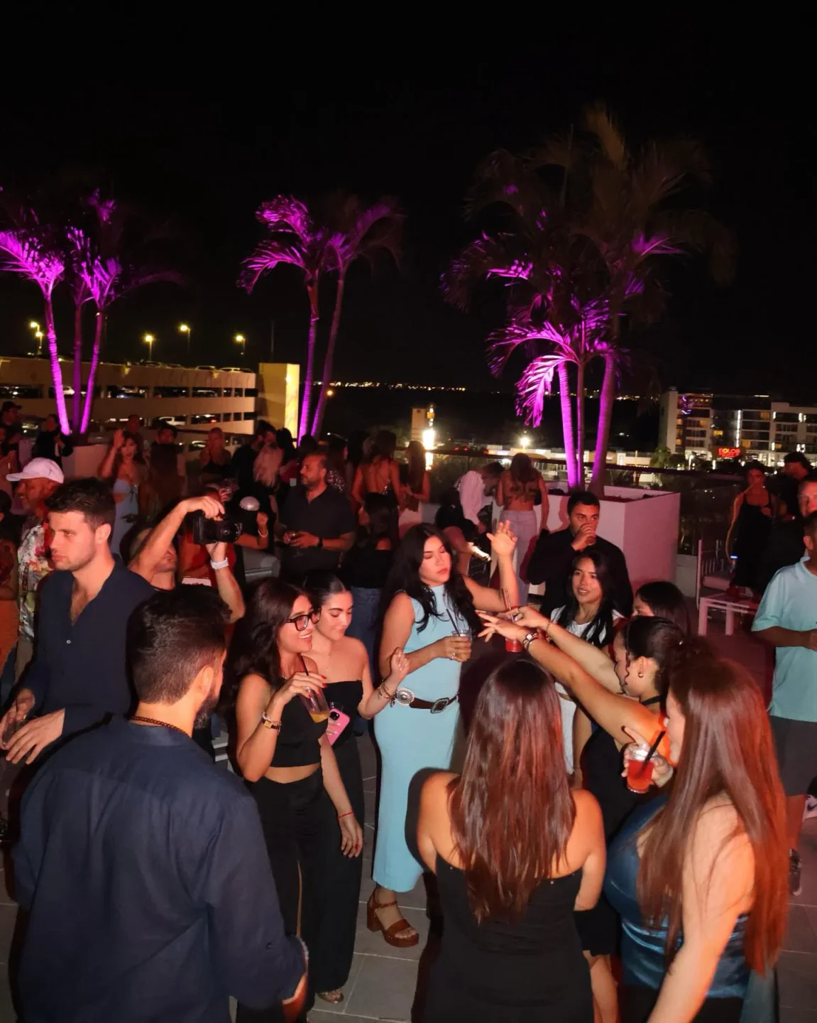 People dancing and enjoying drinks on a vibrant rooftop at Rooftop Live, Miami, under illuminated palm trees.