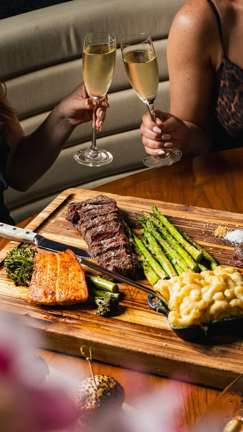 Cheers with champagne at STK restaurant, featuring a steak, salmon, asparagus, and mac & cheese platter.