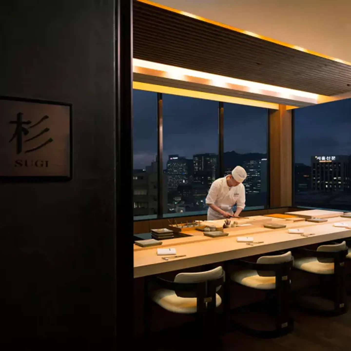 Chef preparing sushi at Sushi Cho in Seoul, with city skyline view at night.
