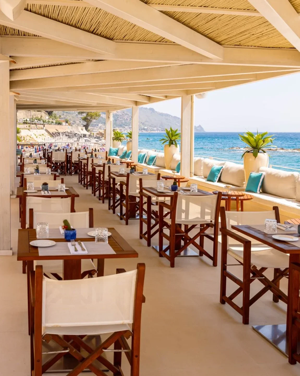 Tao Beach Club Taormina, Sicily, offers stunning sea views from its beachfront dining patio with wooden tables.