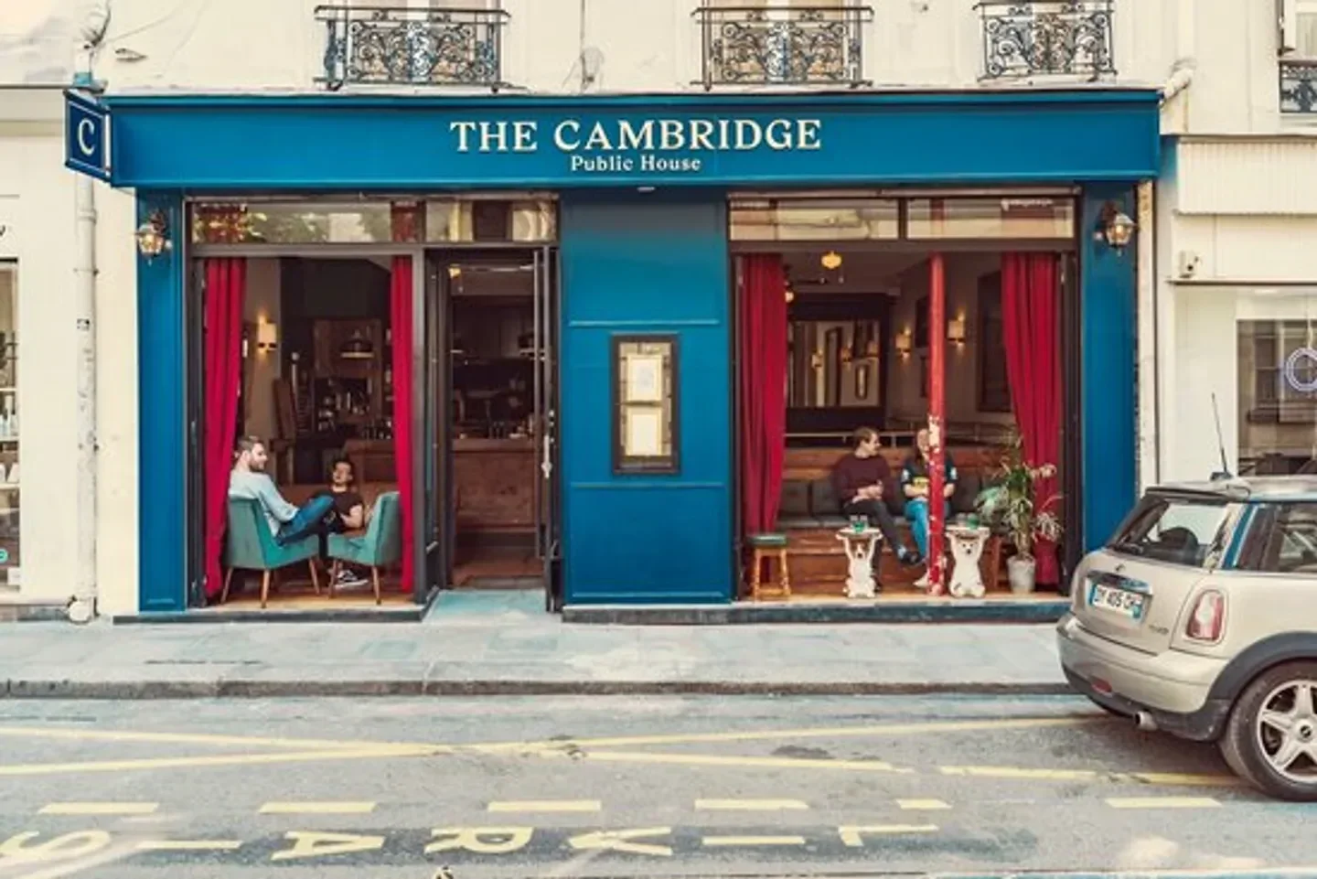 Exterior of The Cambridge Public House in Paris, France, with patrons and car.