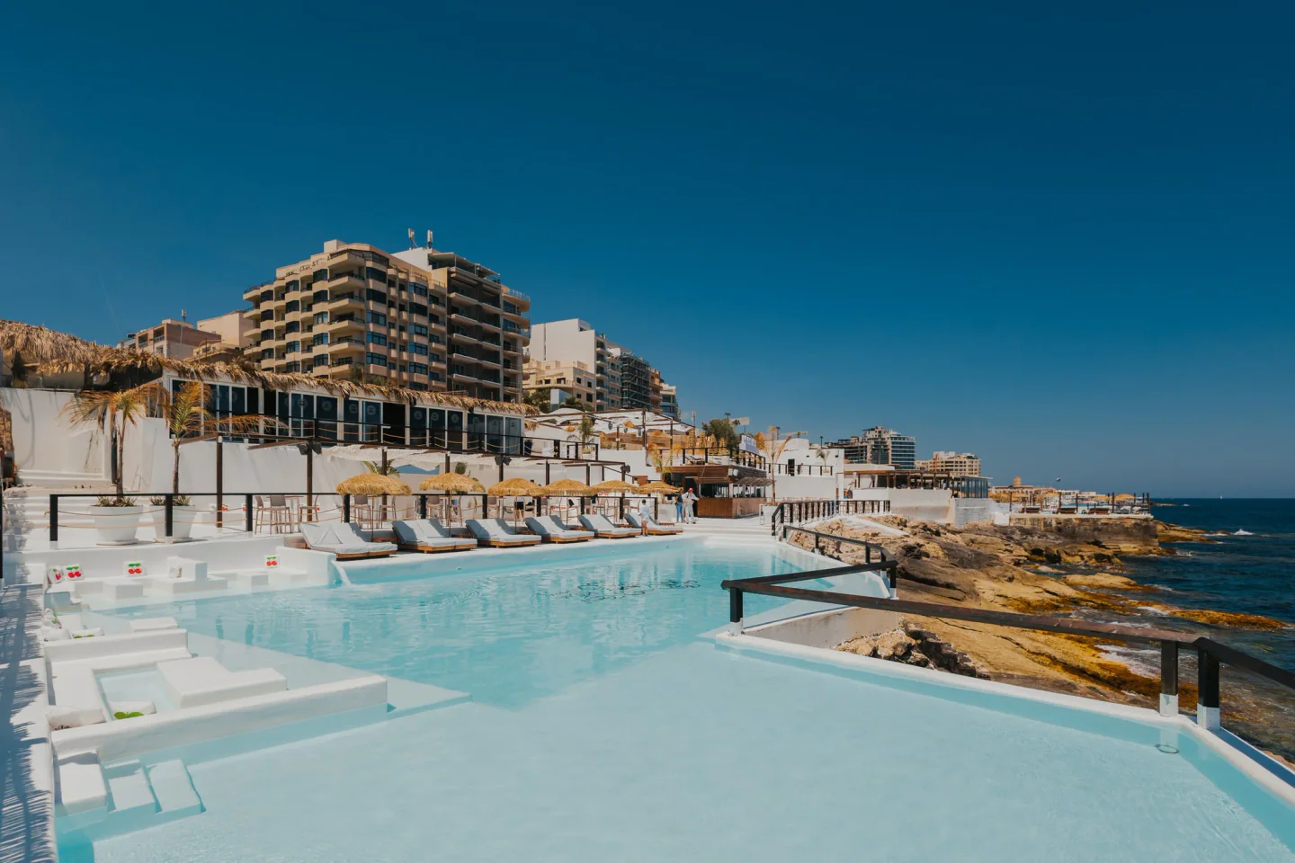 The Toy Room Beach Club in Malta features a stunning white infinity pool overlooking the Mediterranean Sea and rocky coastline.