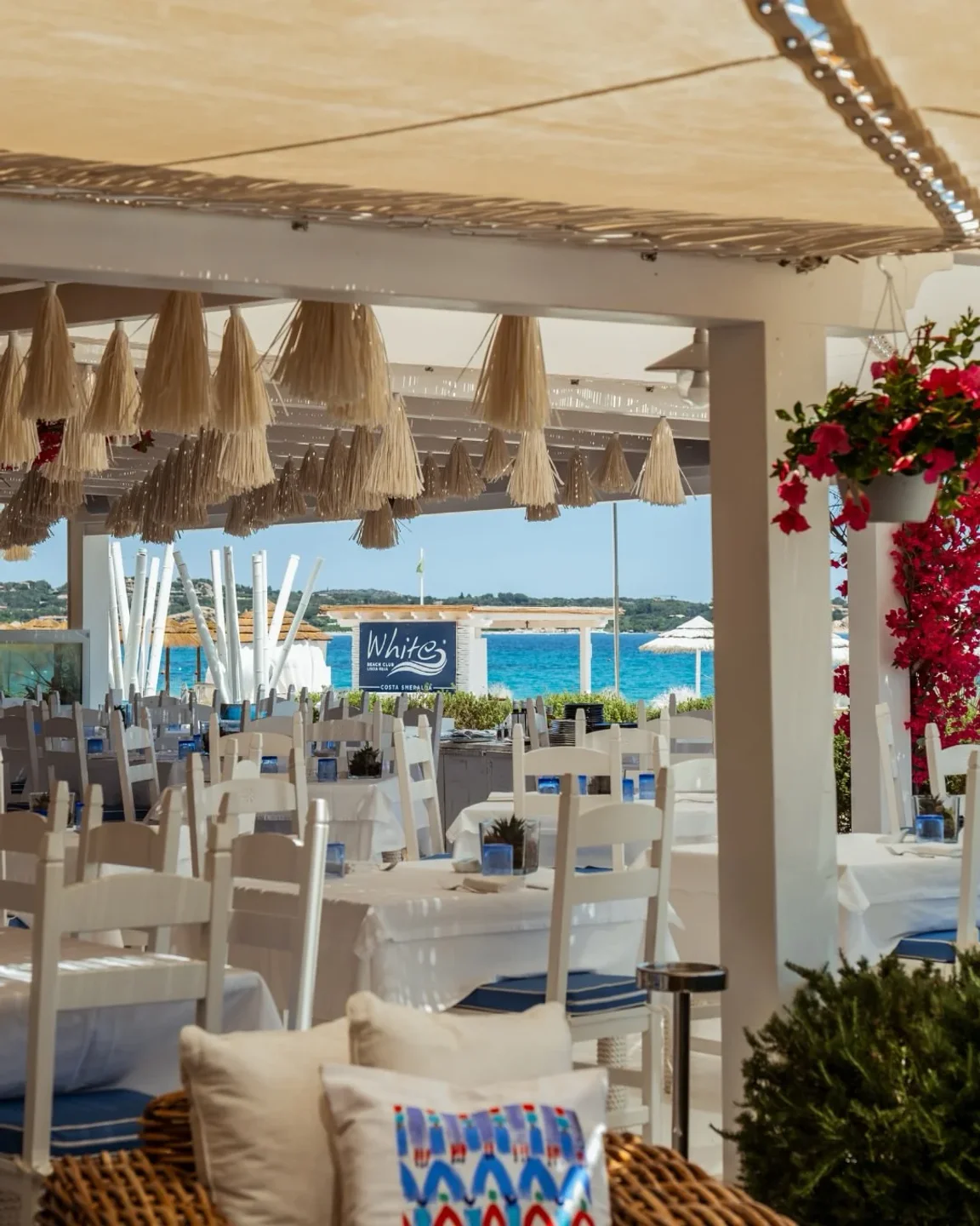 White Beach Club Liscia Ruja seating area with ocean view and vibrant pink flowers.
