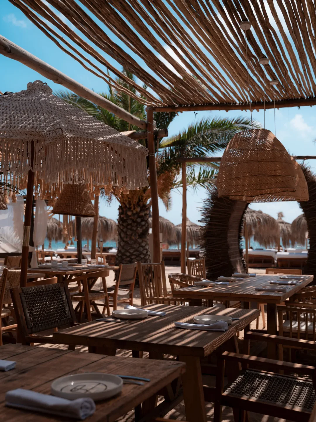Wooden tables and chairs at Zazu beach club in Ibiza, with palm trees and ocean view
